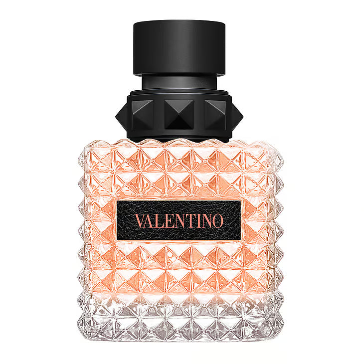 Valentino Donna Born In Roma Coral Fantasy Eau De Parfum – en kvadratisk glasflaska täckt av Valentinos ikoniska nitar. Flaskan innehåller en lysande korallfärgad vätska, och har en svart läderetikett med "VALENTINO" skrivet i korallrosa, stående mot en vit bakgrund.
